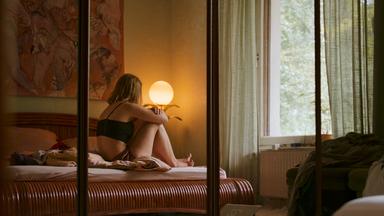 Man sieht einen großen Spiegelschrank, in der Spiegelung erkennt man eine Frau in Unterwäsche auf dem Bett sitzen, sie ist dem Betrachtenden abgewandt. 