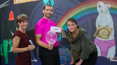 Das Bild zeigt drei Personen, die auf einer Bühne stehen, während im Hintergrund ein farbenfrohes, grafisches Set zu sehen ist.   1. **Personen**:    - Auf der linken Seite steht eine Frau mit kurzen, braunen Haaren. Sie trägt ein dunkelrotes Kleid und hält einen kleinen Gegenstand in der Hand.    - In der Mitte steht ein Mann in einem pinken Shirt und schwarzen Hosen. Er hält einen Luftballon in der einen Hand und ein Blatt Papier in der anderen.    - Die dritte Person, rechts, ist eine Frau mit kurzen, grauen Haaren. Sie trägt einen grauen Pullover und schaut freundlich in die Kamera.  2. **Hintergrund**:     - Der Hintergrund ist bunt gestaltet mit verschiedenen Elementen:       - Eine Regenbogen-Grafik.      - Eine Illustration einer Ratte, die eine Verpackung mit einem Kondom hält.      - Verschiedene fiktive, illustrative Objekte, wie einen Kaktus und bunte Lichtquellen.  Die Szene vermittelt eine humorvolle und informative Atmosphäre, die typisch für eine Wissenschaftssendung ist.