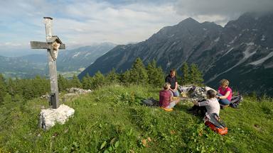 Andrea Ballschuh und eine Wandergruppe sitzen im Grünen neben einem Bergkreuz, im Hintergrund Berge.
