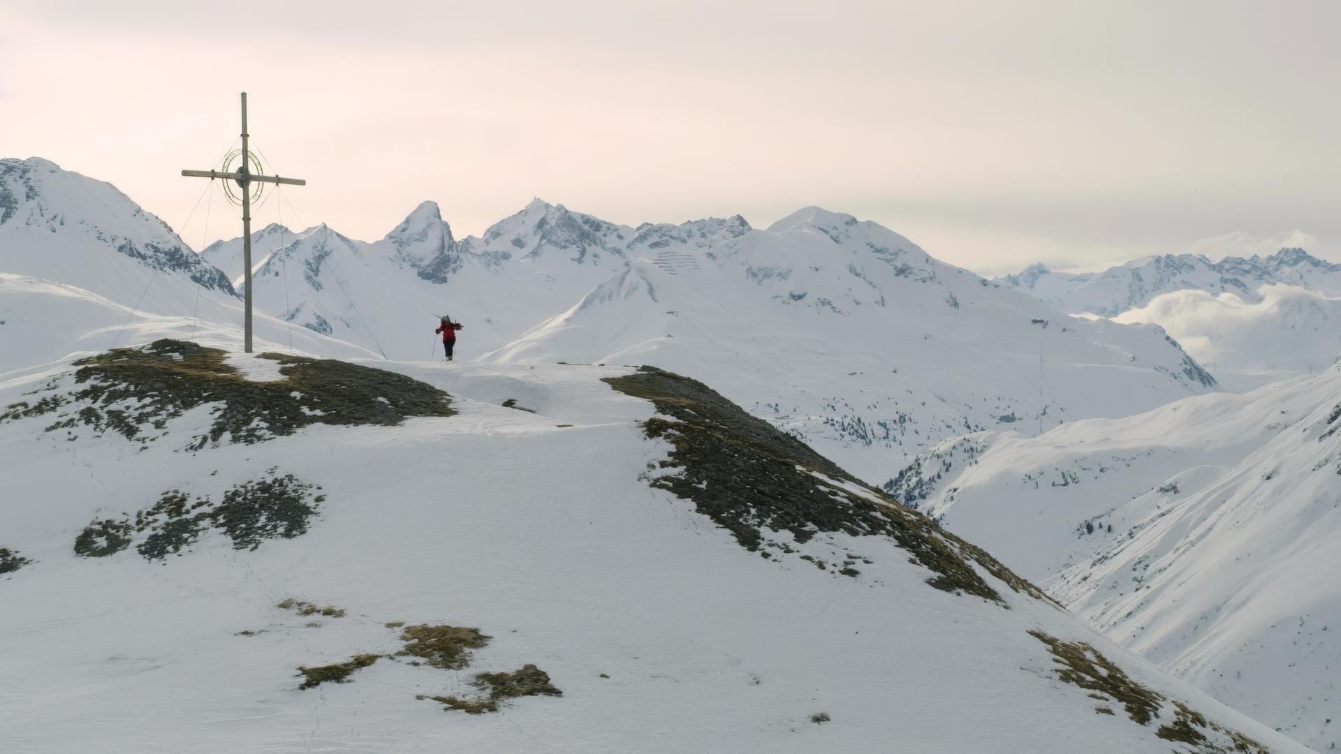 Das Bild zeigt eine schneebedeckte Berglandschaft mit mehreren Gipfeln, die im Hintergrund sichtbar sind. Im Vordergrund steht ein großes Holz-Gipfelkreuz, das von mehreren Seilen gehalten wird. Ein einzelner Mensch, der rot gekleidete Wanderer, geht auf einem schmalen Weg in Richtung des Gipfelkreuzes. Die Szenerie ist von einer ruhigen und majestätischen Atmosphäre geprägt, die die Erhabenheit der Berglandschaft betont. Der Himmel ist in sanften Grautönen gehalten, und die Wolken sind nur leicht sichtbar. Es handelt sich um eine winterliche Umgebung, in der Schnee die Landschaft dominiert.