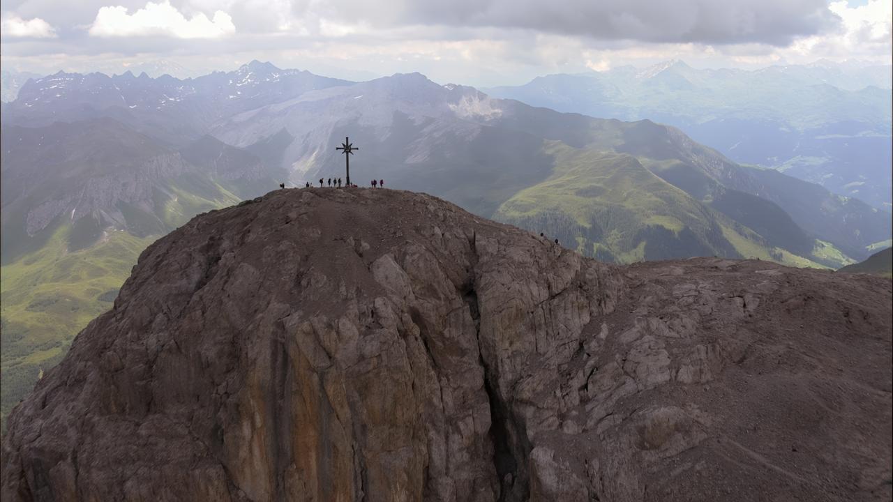 Das Bild zeigt einen höchsten Punkt eines Berges mit einem markanten Gipfelkreuz. Das Kreuz steht auf einer flachen, steinigen Fläche, umgeben von steilen Felswänden und einer zerklüfteten Landschaft. Im Hintergrund erstrecken sich grüne Täler und weitere Bergketten, die sanft in die Ferne verlaufen. Der Himmel ist bewölkt, was eine dramatische Atmosphäre schafft. Am Gipfel sind mehrere Menschen zu sehen, die sich versammelt haben, um die Aussicht zu genießen. Die Szenerie vermittelt ein Gefühl von Erhabenheit und Naturverbundenheit.