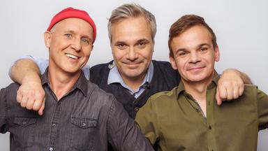 Drei Männer stehen nebeneinander. Sie haken sich mit ihren Armen ein.