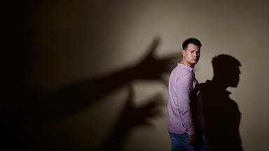 Max Leon steht in Jeans und rosa Hemd seitlich vor einer Wand. Hinter ihm ist sein Schatten an der Wand und Schattenhände, die nach ihm greifen wollen.