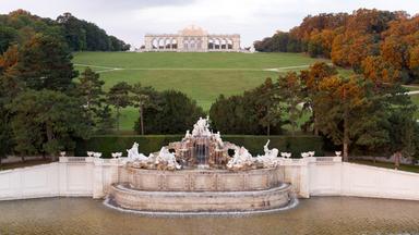 Blick auf die Gloriette im Schlosspark Schönbrunn in Wien, im Vordergrund der Neptunbrunnen mit barocken Skulpturen, im Hintergrund das monumentale Bauwerk auf einem Hügel.