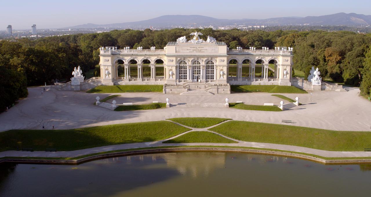 Frontalansicht der Gloriette im Schlosspark Schönbrunn in Wien, dahinter bewaldete Hügel und die Stadt im Hintergrund, vorne der künstlich angelegte Teich.