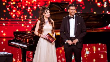 Eine Frau und ein Mann in Abendrobe stehen auf einer rot beleuchteten Bühne mit einem Flügel im Hintergrund