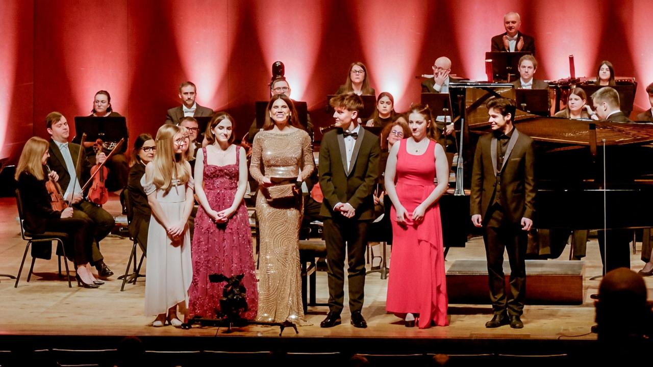 Sechs Personen stehen vor einem Orchester auf einer Konzertbühne in festlicher Abendkleidung, im Hintergrund sitzen Musikerinnen und Musiker mit Instrumenten.