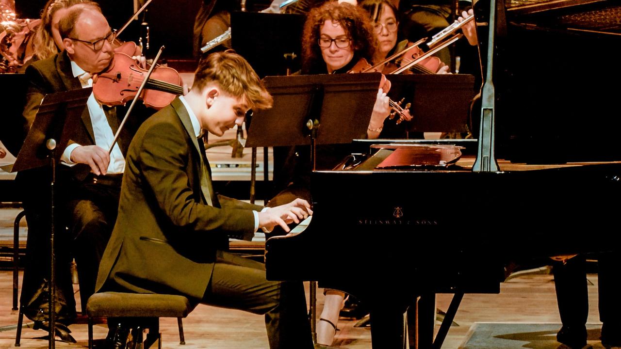 Ein junger Mann im Anzug spielt konzentriert auf einem Steinway-Flügel, hinter ihm sitzt ein Orchester mit Streichinstrumenten.