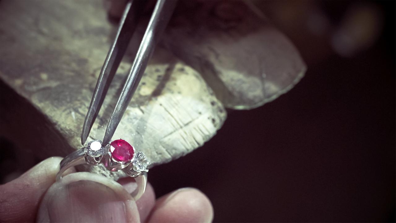 Das Bild zeigt eine Nahaufnahme, in der eine Hand mit einem silbernen Ring zu sehen ist. Der Ring ist mit einem roten Stein und zwei weißen Steinen verziert, die deutlich sichtbar sind. In der Hand wird eine Zange gehalten, mit der offenbar ein Stein in den Ring eingesetzt wird. Der Hintergrund ist dunkel und unscharf, wodurch der Fokus auf dem Ring und den handwerklichen Arbeiten liegt. Unter der Hand befindet sich eine Werkbank aus Holz, auf der der Ring platziert ist. Die Oberfläche der Werkbank zeigt Gebrauchsspuren und einen natürlichen, rustikalen Look. Der gesamte Eindruck vermittelt die Atmosphäre eines Goldschmiedewerkstatt, in der sorgfältige Handarbeit zur Schmuckherstellung durchgeführt wird.