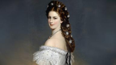 Das Bild zeigt eine Frau in einem historischen Kleid, das elegant und prächtig aussieht. Sie hat lange, leicht gewellte, brünette Haare, in denen mehrere glitzernde Sternenornamente eingeflochten sind. Ihr Gesicht ist sanft ausdrucksvoll, mit einem leichten Lächeln, das einen freundlichen Eindruck vermittelt. Die Frau trägt eine perlenbesetzte Halskette. Ihr Kleid hat weite, dekorierte Ärmel und ist bis zur Schulter ausgeschnitten. Der Hintergrund ist neutral gehalten, sodass der Fokus auf der Frau und ihrem Schmuck liegt.   Das Bild vermittelt eine Atmosphäre von Eleganz und Anmut. Es ist ein Porträt, das sowohl Kleidung als auch Kunstfertigkeit im Goldschmiedehandwerk thematisiert.