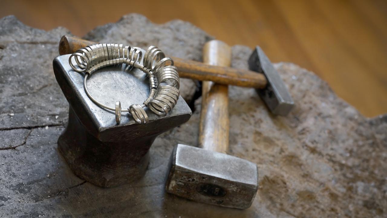 Das Bild zeigt eine Werkstatt-Szene, die dem handwerklichen Prozess des Goldschmiedens gewidmet ist. Im Vordergrund steht ein Amboss aus Metall, auf dem mehrere kleine runde Metallringe abgelegt sind. Diese Ringe sind möglicherweise Prototypen für zukünftigen Schmuck.   Neben dem Amboss befinden sich zwei Hämmer, einer mit einem schweren Kopf aus Metall und der andere mit einem Holzgriff, auf einer steinernen Unterlage. Die Umgebung wirkt rustikal, mit einem sichtbaren Holzboden und unscharfen Hintergrunddetails, die möglicherweise Werkzeuge oder weitere Materialien zeigen.  Das Bild vermittelt einen handwerklichen Charakter und zeigt die grundlegenden Werkzeuge, die Gold- und Silberschmiede in ihrer Arbeit verwenden.