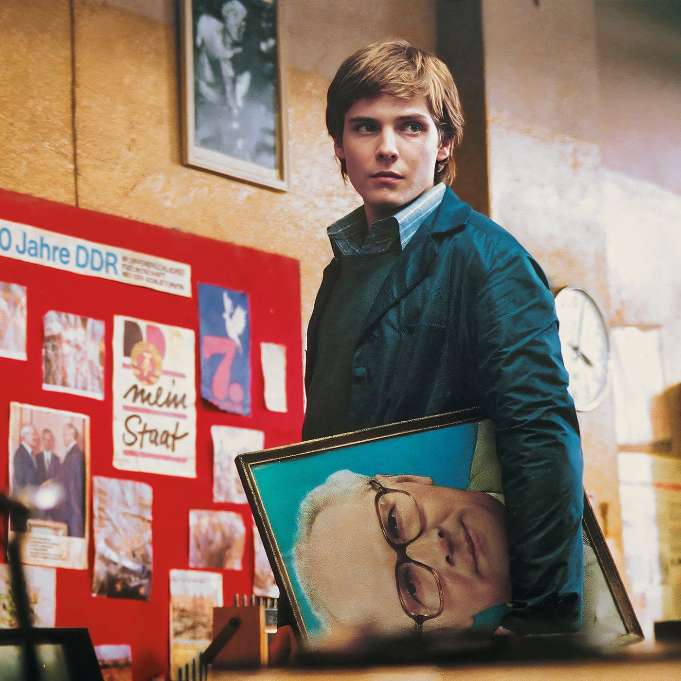 Alex (Daniel Brühl) blickt in einer Art Lagerhalle suchend um sich. Unter dem linken Arm trägt er ein Porträt Erich Honeckers. Im Hintergrund sieht man eine Fototafel mit der Überschrift „40 Jahre DDR“.