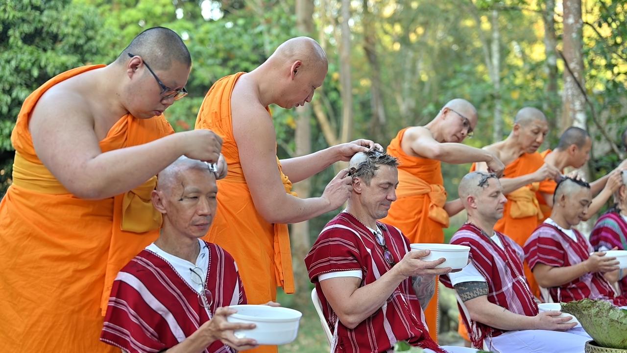 Das Bild zeigt eine Szene in einem buddhistischen Kloster in Thailand. Im Vordergrund sitzen mehrere Männer, die jeweils eine Schüssel in den Händen halten. Diese Männer tragen traditionelle, gestreifte Kleidung in Rot und Weiß. Ihre Köpfe sind teilweise rasiert, während einige von ihnen noch längeres Haar haben.   Hinter ihnen stehen mehrere Mönche in orangefarbenen Roben. Diese Mönche führen eine zeremonielle Kopfrasur durch, was ein wichtiger Teil der Ordination in der buddhistischen Tradition ist. Die Mönche scheinen konzentriert zu arbeiten, während sie die Haarpracht der Männer, die für diese Zeremonie teilnehmen, schneiden.   Die Umgebung ist grün und naturnah, mit vielen Bäumen und Pflanzen im Hintergrund, die eine friedliche und spirituelle Atmosphäre schaffen. Es gibt keine Hinweise auf moderne Technologie oder Ablenkungen, was die spirituelle Konzentration der Szene unterstreicht.   Die gesamte Darstellung vermittelt ein Gefühl von Gemeinschaft, Tradition und der Suche nach innerem Frieden.
