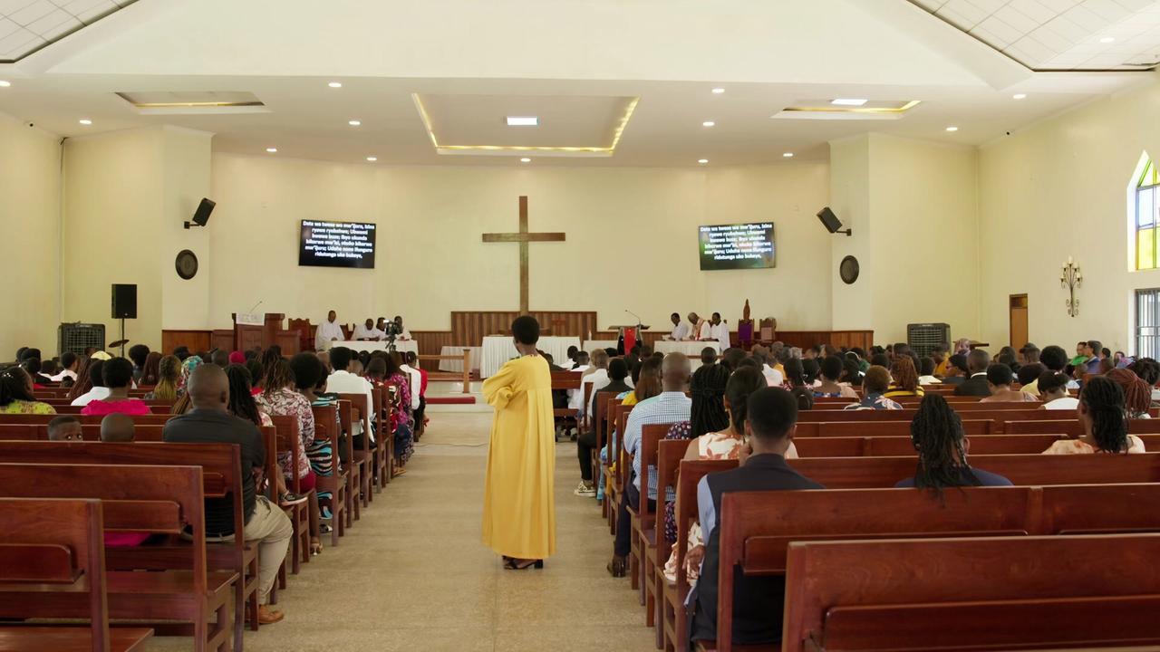 Das Bild zeigt das Innere einer Kirche in Ruanda während einer Messe. Der Raum ist hell erleuchtet, mit einem hohen, schrägen Dach. An der Wand hinter einem Altar befindet sich ein großes, einfaches Holzkreuz. Vor dem Altar stehen mehrere Personen in weißen Gewändern, die anscheinend eine religiöse Zeremonie durchführen.  Die Bankreihen vor dem Altar sind nahezu vollständig besetzt. Viele Gläubige sitzen in hölzernen Stühlen, die in Reihen angeordnet sind. Die Anwesenden sind überwiegend in farbenfrohe Kleidungsstücke gehüllt, die sowohl traditionelle als auch moderne Stile zeigen. Einige Menschen halten Broschüren oder schauen auf Bildschirme, die an den Wänden hängen und möglicherweise die Liedtexte oder Gebete anzeigen.  Im Vordergrund ist eine Frau in einem gelben Kleid zu sehen, die sich in Richtung Altar bewegt. Der Raum hat einen klaren akustischen Charakter, ideal für Gesänge und Predigten.  Insgesamt vermittelt die Szene eine spirituelle Atmosphäre, geprägt von Gemeinschaft und Gottesdienst.