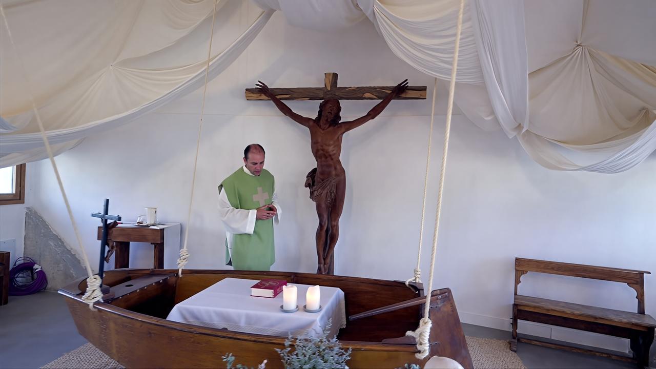 Das Bild zeigt einen kleinen, hellen Raum, der mit einem weißen Stoffdach dekoriert ist, das sanft herunterhängt. In der Mitte des Raumes steht ein hölzernes Boot, das als Altar fungiert. Auf dem Altar liegen eine weiße Tischdecke und eine rote Bibel. Vor dem Boot stehen zwei brennende Kerzen.  An der Wand hinter dem Boot hängt ein großer, geschnitzter Kreuzweg mit einer Statue von Jesus, der mit weit ausgebreiteten Armen da steht. Der Raum ist schlicht und minimalistisch eingerichtet. Zu sehen sind auch eine kleine, dunkle Holzbank sowie ein Regal mit einigen religiösen Gegenständen.  Eine Person, gekleidet in einem grünen Gewand mit weißen Ärmeln, steht am Altar und scheint zu beten. Der Raum vermittelt eine Atmosphäre der Ruhe und Besinnung.