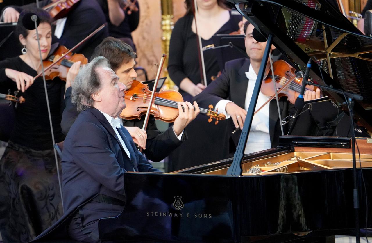 Das Bild zeigt eine Live-Aufführung bei einem Musikfestival. Im Vordergrund sitzt ein Pianist an einem schwarzen Flügel, der offen steht, sodass die Klaviatur und einige der inneren Mechanismen sichtbar sind. Der Pianist hat graue Haare und trägt einen schwarzen Anzug mit einer weißen Fliege.   Hinter ihm befindet sich ein Orchester mit mehreren Musikern, darunter Geiger, die ihre Instrumente spielen. Einige der Orchestermusiker sind in Schwarz gekleidet. Die Szenerie wirkt festlich und ist Teil des Grafenegg Festivals in Österreich, einem bedeutenden Musikereignis. Im Hintergrund sind möglicherweise architektonische Elemente zu erkennen, die auf die malerische Kulisse hinweisen, die das Festival umgibt.   Das Licht ist hell und variiert zwischen warmen und kühlen Tönen, was zur Atmosphäre des Konzerts beiträgt.