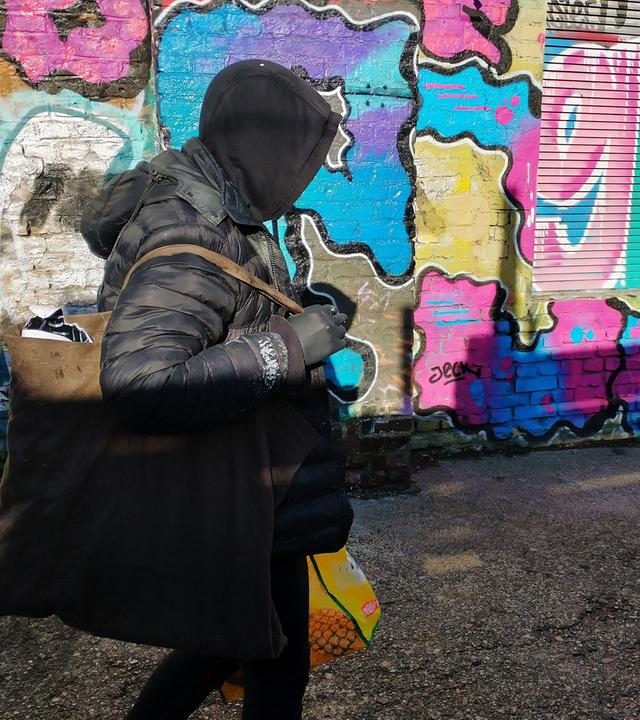 Das Bild zeigt einen Menschen, der schwarz gekleidet ist und eine Kapuze trägt. Man sieht sein Gesicht nicht. In den Händen, die mit schwarzen Handschuhen versteckt sind, trägt er größere Tüten. Er läuft an einer Wand entlang, die voll von bunten Graffitis ist.