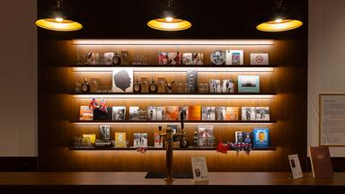 Eine Theke mit Zapfhahn und dahinter ist ein Bücherregal, in dem verschiedene Bilder, Bücher, Parfums und auch Flaggen stehen. Von oben hängen drei Lampen runter ins Bild.