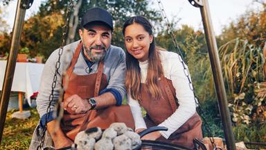 Ein Mann und eine Frau blicken in die Kamera. Vor ihnen befindet sich ein mit Kohlen bedeckter Gusseisentopf auf einem Dreibein-Rost.