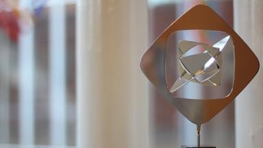 Das Bild zeigt eine moderne, abstrakte Skulptur aus poliertem Metall, die auf einem schwarzen Sockel steht, vor einem unscharfen Hintergrund eines Fensters mit weißen Vertikalstreben. 