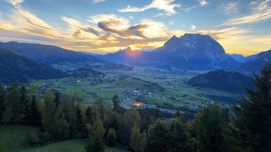 Blick ins Ennstal bei Sonnenuntergang mit dem markanten Grimming im Hintergrund; darunter verstreute Ortschaften, Felder und bewaldete Hügel.