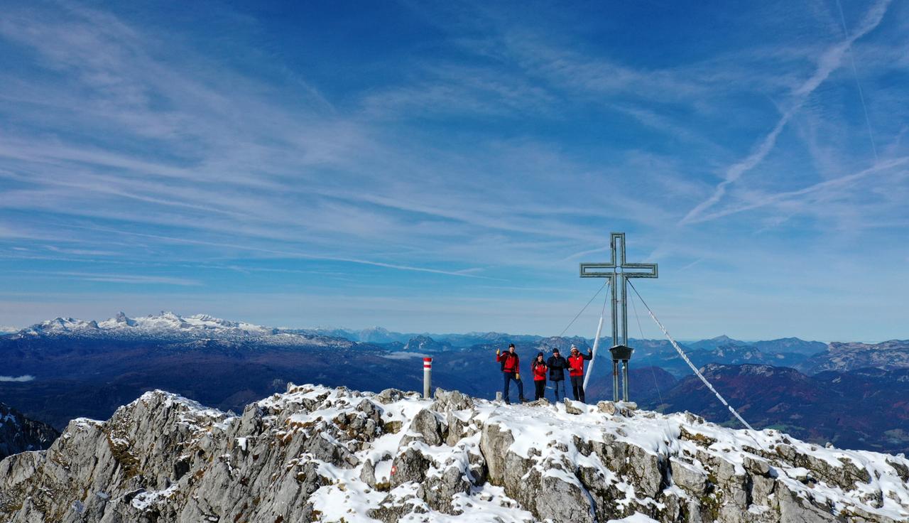 Fünf Bergsteiger stehen auf einem schneebedeckten Gipfel neben einem großen Gipfelkreuz mit Weitblick über verschneite Alpenketten und einen tiefblauen Himmel.
