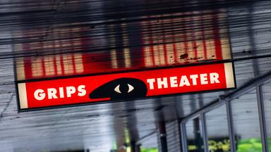 Über dem Eingang eines modernen Gebäudes leuchtet eine rote Neontafel, auf der Grips Theater zu lesen ist.