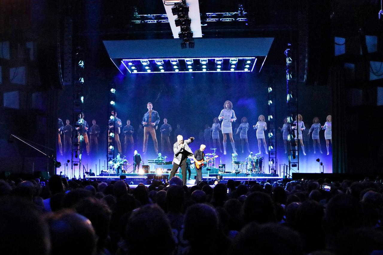 Herbert Grönemeyer singend auf einer blau beleuchteten Bühne. Auf die Leinwand sind Tänzer*innen projiziert.