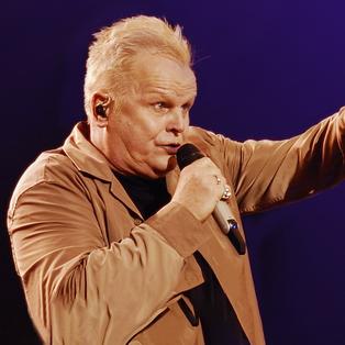 Herbert Grönemeyer singend mit Mikrofon vor dem Mund, den linken Arm mit Zeigefinger deutend zum Publikum. Er trägt einen Mantel in beige.