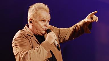 Herbert Grönemeyer singend mit Mikrofon vor dem Mund, den linken Arm mit Zeigefinger deutend zum Publikum. Er trägt einen Mantel in beige.