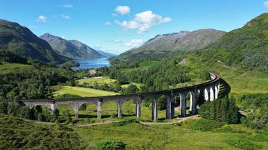 Luftaufnahme einer historischen Eisenbahnbrücke in den schottischen Highlands mit Bergen, Seen und grünen Tälern.