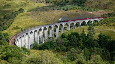 Historischer Dampfzug fährt über ein großes Steinviadukt in den grünen schottischen Highlands.