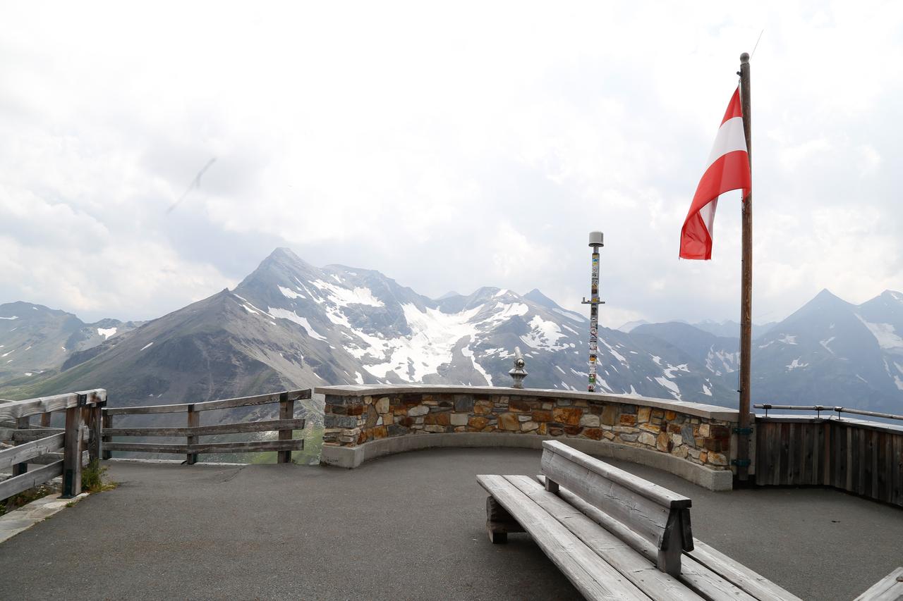 Das Bild zeigt einen Aussichtspunkt in den Alpen, am Ende der Hochalpenstraße auf den Großglockner, dem höchsten Berg Österreichs. Im Vordergrund befindet sich eine geschwungene Mauer aus Bruchstein, die mit einem Holzgeländer ergänzt ist. Darauf steht ein metallisches Objekt, vermutlich eine Wetterstation, das mit Aufklebern verziert ist. Auf der rechten Seite weht eine rot-weiße Fahne, das Nationalflagge Österreichs.  Im Hintergrund sind majestätische Berggipfel zu sehen, einige mit Schnee bedeckt. Der Himmel ist leicht bewölkt, was eine gedämpfte Lichtstimmung erzeugt. Auf dem Bürgersteig liegen einfache Holzbänke, die zum Verweilen einladen. Die gesamte Szenerie vermittelt ein Gefühl von Naturverbundenheit und den Einfluss des Menschen auf die alpine Landschaft.