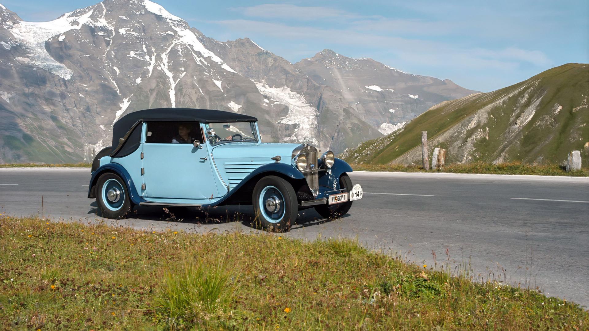 Das Bild zeigt einen historischen Oldtimer, der auf einer gebogenen Straße auf der Großen Glockner Hochalpenstraße steht. Der Wagen ist in einem blassen Blau mit einem schwarzen Faltverdeck lackiert. Im Hintergrund erheben sich majestätische Berge, einige mit schneebedeckten Gipfeln, während andere grüne Hänge aufweisen. Der Himmel ist klar und blau, mit wenigen Wolken. Um den Wagen herum wachsen einige Wiesenblumen, die dem Bild eine lebendige Note verleihen. Die Umgebung strahlt Natur und Ruhe aus, was den Eindruck eines faszinierenden Naturerlebnisses verstärkt. Die Hochalpenstraße ist für ihre landschaftliche Schönheit und die Möglichkeit von Oldtimerfahrten bekannt.