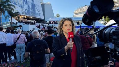Eine Frau mit einem 3sat-Mikrofon wird von einer Kamera gefilmt, während sie vor dem Kino in Cannes steht und einen Aufsager macht.