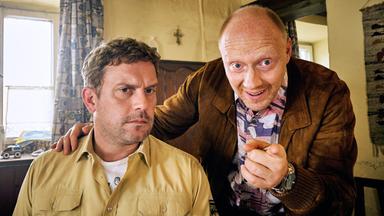 Ein sitzender Mann in Polizeiuniform. Ein weiterer Mann hinter ihm legt ihm die Hand auf die Schulter und deutet in Richtung Kamera.