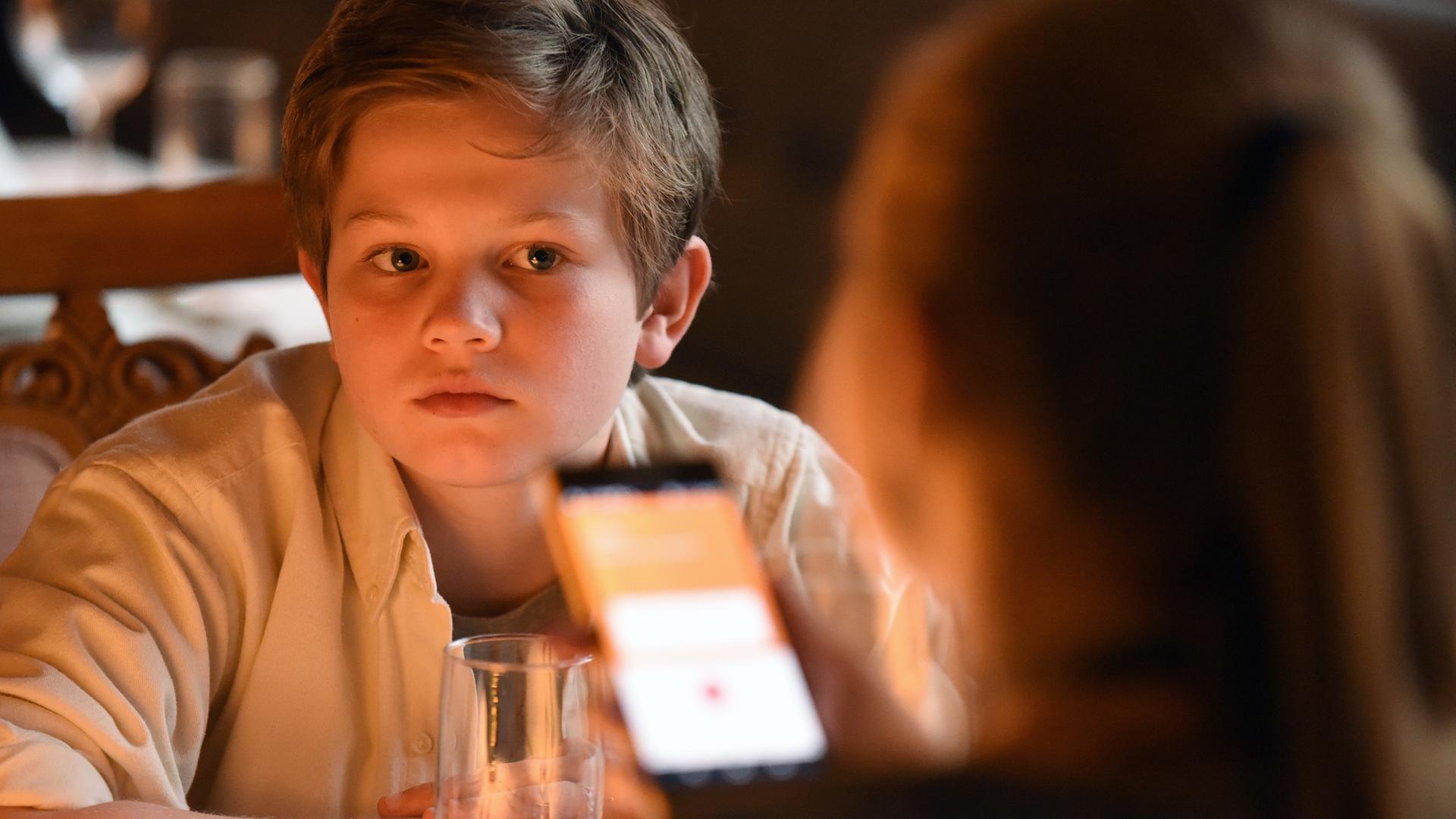 "Frühling - Gute Väter, schlechte Väter": Nicky Söringer (Valentin Wessely) sitzt in einem Restaurant an einem Tisch und blickt erwartungsvoll auf seine Schwester Paula (Lotte Hanné), die auf ihr Handy schaut.