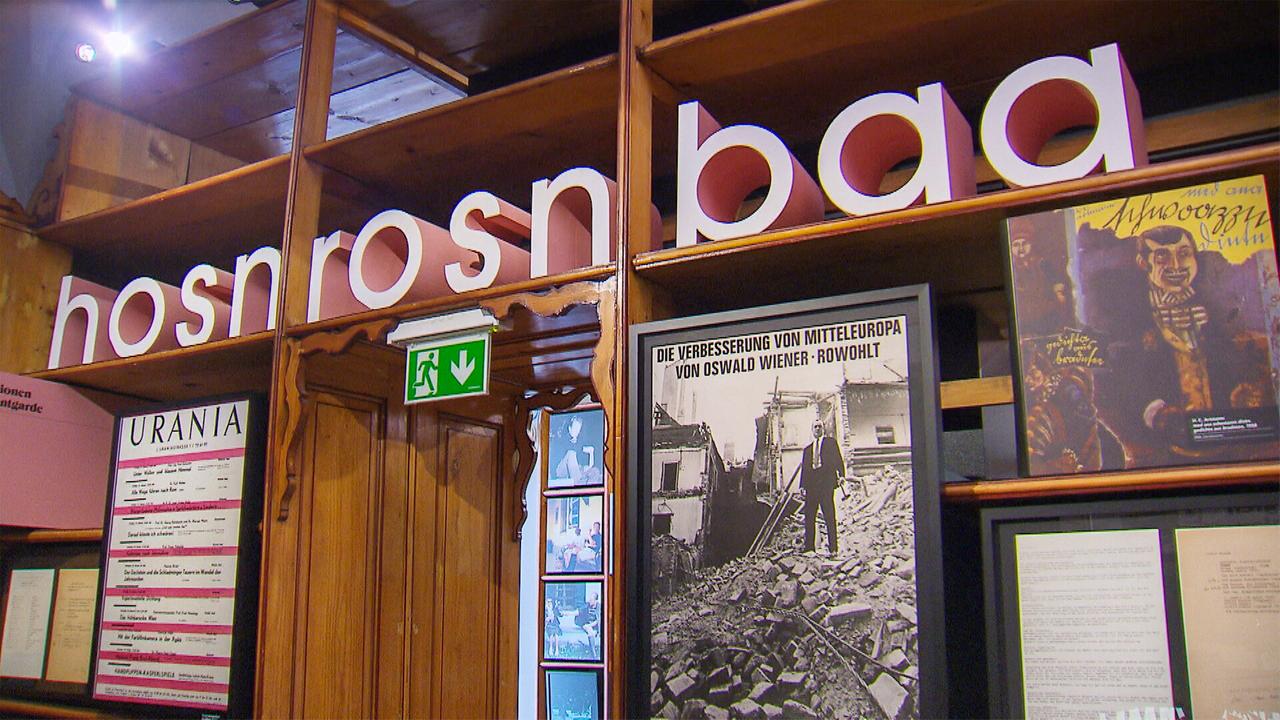 Das Bild zeigt eine Innenansicht des Wiener Literaturmuseums. Im oberen Bereich ist das Wort "hosnrosnbaa" in großen, bunten Buchstaben zu sehen. Darunter befinden sich mehrere Rahmen mit verschiedenen Informationen und Bildern. Ein auffälliges Element ist ein Plakat, das den Titel "Die Verbesserung von Mitteleuropa" von Oswald Wiener zeigt, daneben eine weitere Darstellung. Im Hintergrund erkennt man eine Holzwand mit Regalbrettern, die einige Ausstellungsstücke und Informationsblätter beherbergen. Zudem ist ein Notausgangsschild sichtbar. Die gesamte Atmosphäre vermittelt einen literarischen und kulturellen Eindruck, passend zur Thematik des Museums.