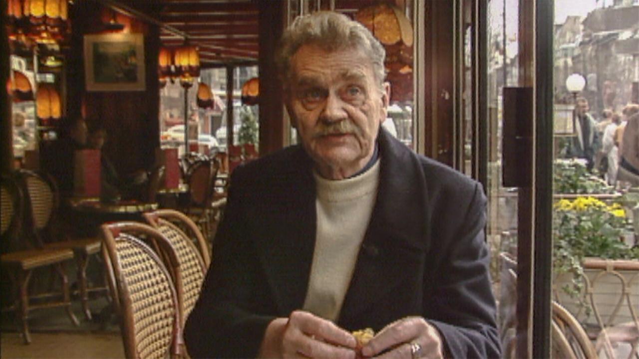 Das Bild zeigt einen älteren Mann, der in einem Café sitzt. Er hat graue Haare und einen Schnurrbart. Er trägt einen schwarzen Mantel und einen hellen Pullover darunter. Seine Hände sind sichtbar, während er etwas in der linken Hand hält. Im Hintergrund sind die charakteristischen Möbel eines Cafés zu sehen, einschließlich geflochtener Stühle und runder Tische. Die Beleuchtung ist warm und einladend, mit mehreren pendelnden Lampen, die an der Decke befestigt sind. Durch die Fenster sind verschwommene äußere Ansichten der Stadt zu erkennen, einschließlich Pflanzen und Passanten.