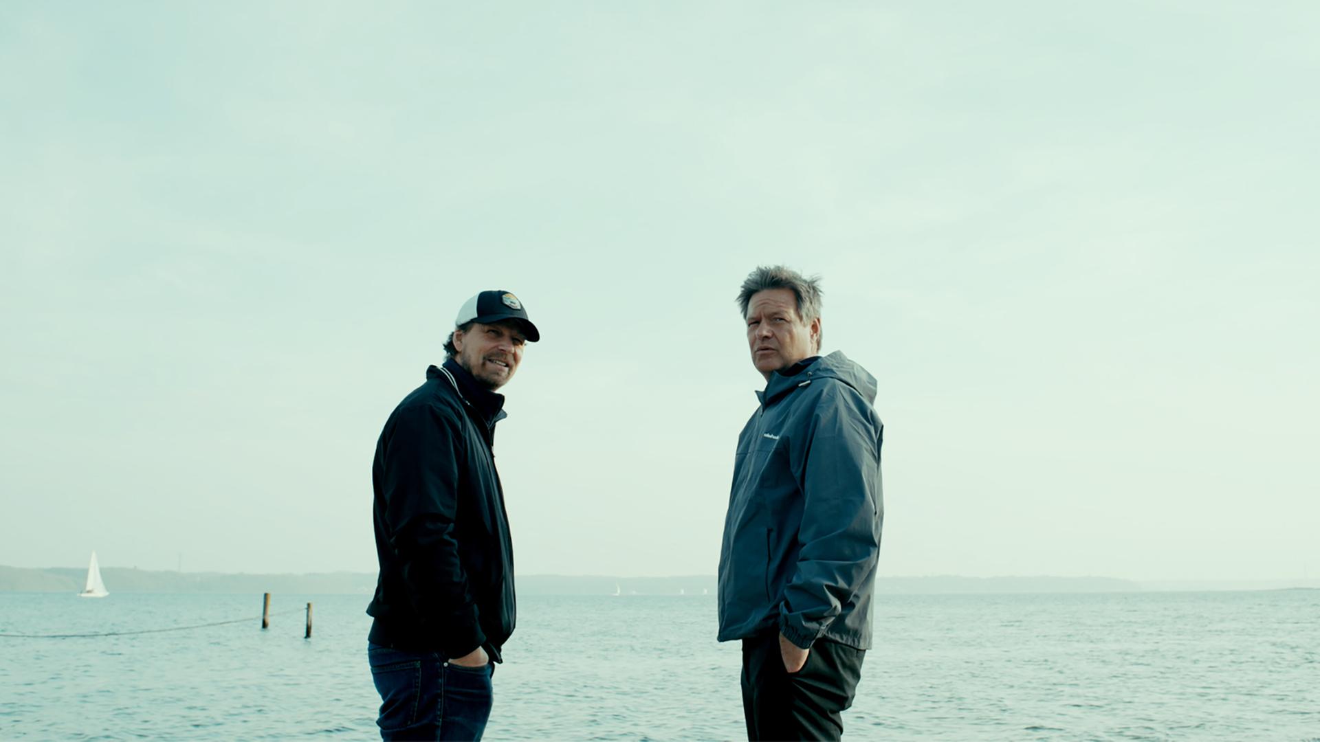 Zwei Männer in Windbreakern stehen an einem Strand.