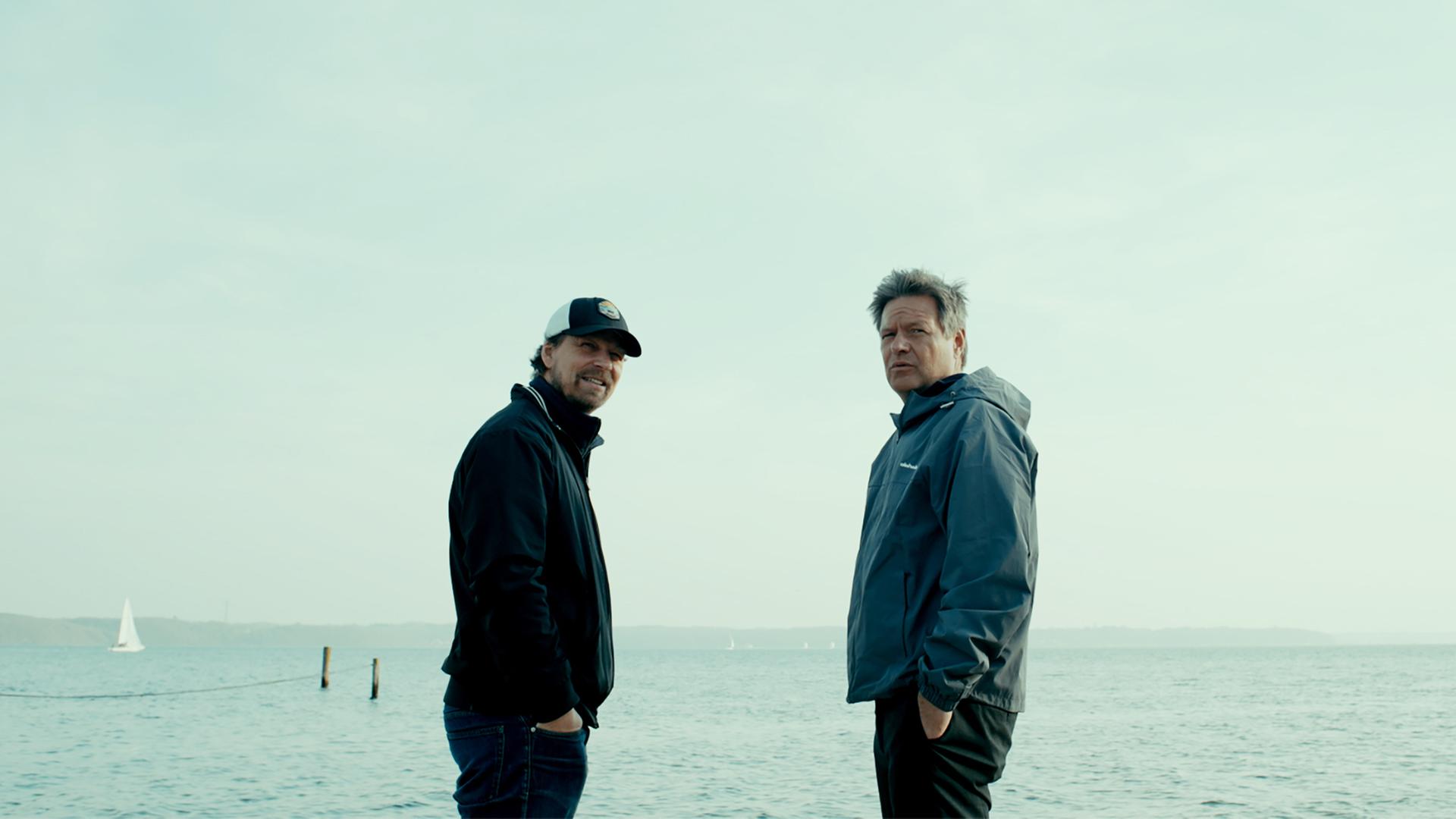 Zwei Männer in Windbreakern stehen an einem Strand.