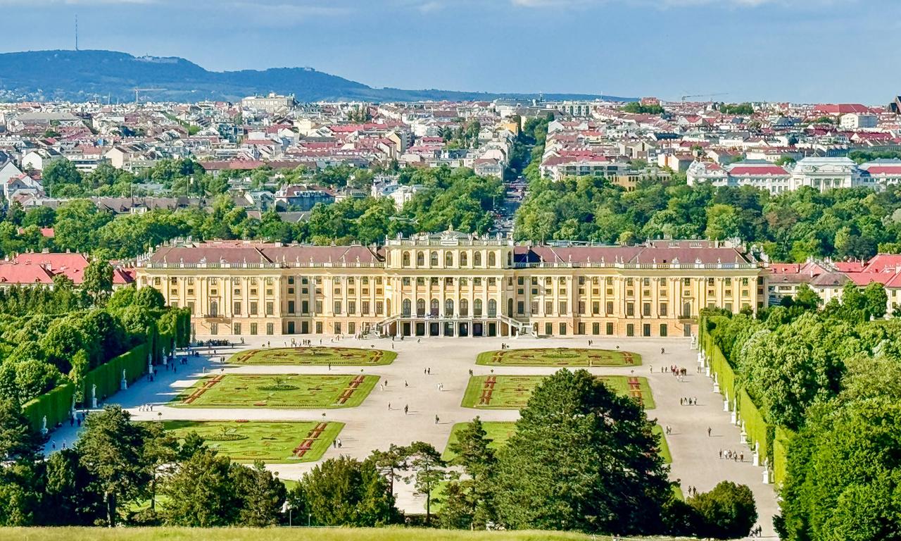 Das Bild zeigt das Schloss Schönbrunn in Wien, Österreich. Im Vordergrund ist die imposante Fassade des Schlosses sichtbar, das in einem hellen Gelbton gestrichen ist und mit einer Vielzahl von Fenstern und Kunstwerken geschmückt ist. Vor dem Schloss erstreckt sich ein weitläufiger Park mit geometrisch angelegten Gartenanlagen, die kunstvoll mit Blumenbeeten und Hecken gestaltet sind.  Rund um das Schloss befinden sich Spazierwege, auf denen mehrere Personen flanieren, was die lebendige Atmosphäre des Ortes unterstreicht. Im Hintergrund erhebt sich eine Stadtlandschaft mit verschiedenen Wohngebäuden, deren Dächer in verschiedenen Rottönen gedeckt sind. Der Himmel ist klar und blau, was den Eindruck eines sonnigen Tages vermittelt.  In der Ferne sind sanfte Hügel oder ein Gebirge zu erkennen, was dem Bild zusätzliche Tiefe und Dimension verleiht. Die Szene vermittelt den Eindruck von historischer Pracht und natürlicher Schönheit, typischen Merkmalen der Umgebung von Schönbrunn.