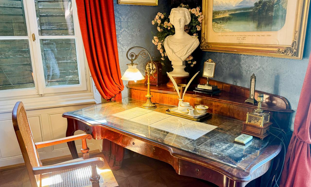 Das Bild zeigt das Arbeitszimmer von Kaiser Franz Joseph I. in Bad Ischl, Oberösterreich. Der Raum ist elegant und historisch gestaltet. Im Vordergrund steht ein großer, geschwungener Schreibtisch aus Holz, dessen Oberfläche mit einer glasartigen Abdeckung versehen ist. Auf dem Tisch liegen mehrere Dokumente, die handschriftliche Texte zeigen. Zu den prominentesten Gegenständen gehören eine dekorative Lampe mit einer opalenen Schirm, eine kleine figurative Statue (eine Büste) und eine feine Teekanne. An der linken Seite des Bildes sieht man eine Holzbank mit einer geflochtenen Sitzfläche. Im Hintergrund hängen rote Vorhänge, die einen Kontrast zu den blauen Wänden bilden.   An der Wand ist ein gerahmtes Bild sichtbar, das eine Landschaft darstellt, und eine kleine Uhr steht ebenfalls auf dem Tisch. Die Gesamtharmonie des Raums vermittelt einen Eindruck von imperialem Glanz und historischem Erbe.