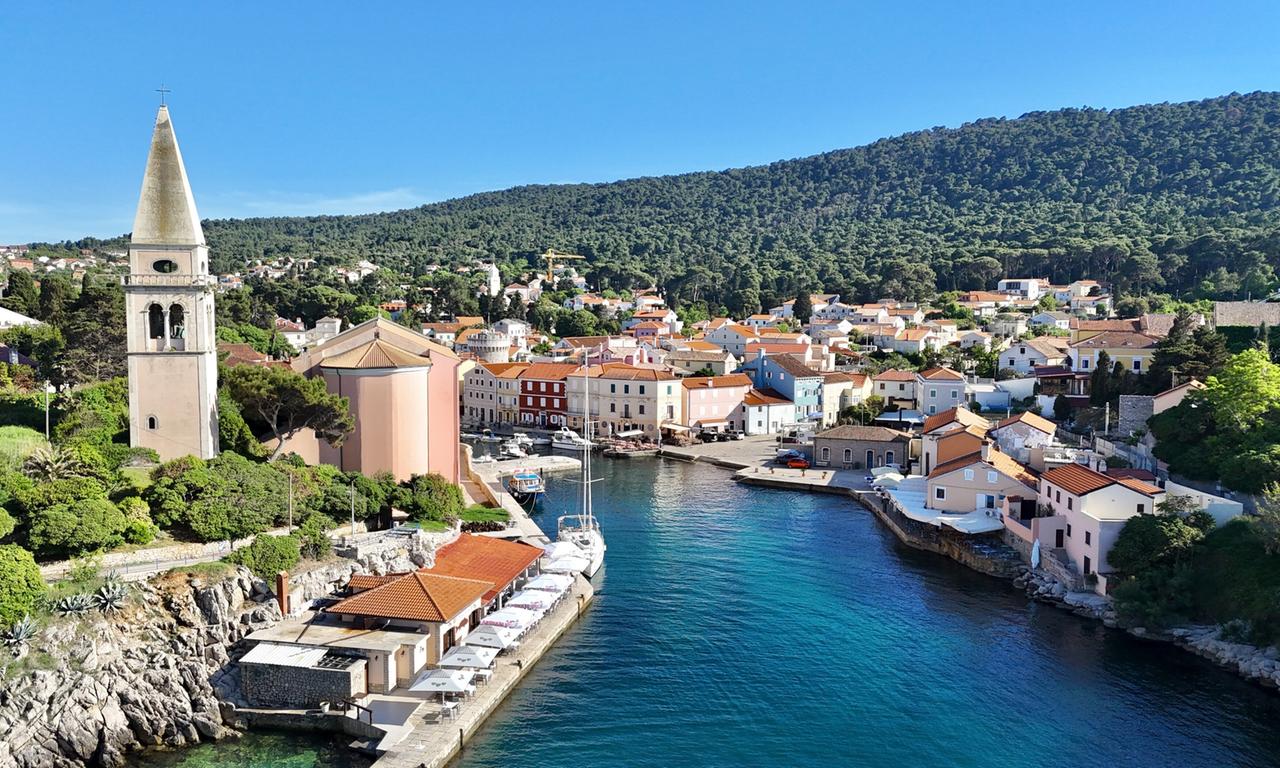 Das Bild zeigt den Küstenort Veli Lošinj in Kroatien. Im Vordergrund fließt ein ruhiger, blauer Hafen mit mehreren Booten, die dort vertäut sind. Auf der linken Seite des Bildes steht ein hoher, spitzer Kirchturm, der aus hellem Stein gebaut ist. Direkt daneben befindet sich ein Gebäude mit rosa Fassade und einem mehrseitigen Dach. Die Küste ist von üppigem Grün umgeben, das in verschiedenen Brauntönen und Grüntönen schimmert. Im Hintergrund sind weitere Häuser zu sehen, die in verschiedenen Farben, darunter Gelb, Rot und Blau, gestaltet sind. Hinter den Häusern erheben sich bewaldete Hügel, die den Ort umrahmen. Der Himmel ist klar und blau, was auf einen sonnigen Tag hinweist. Insgesamt vermittelt das Bild eine ruhige, malerische Küstenatmosphäre, die typisch für mediterrane Orte ist.