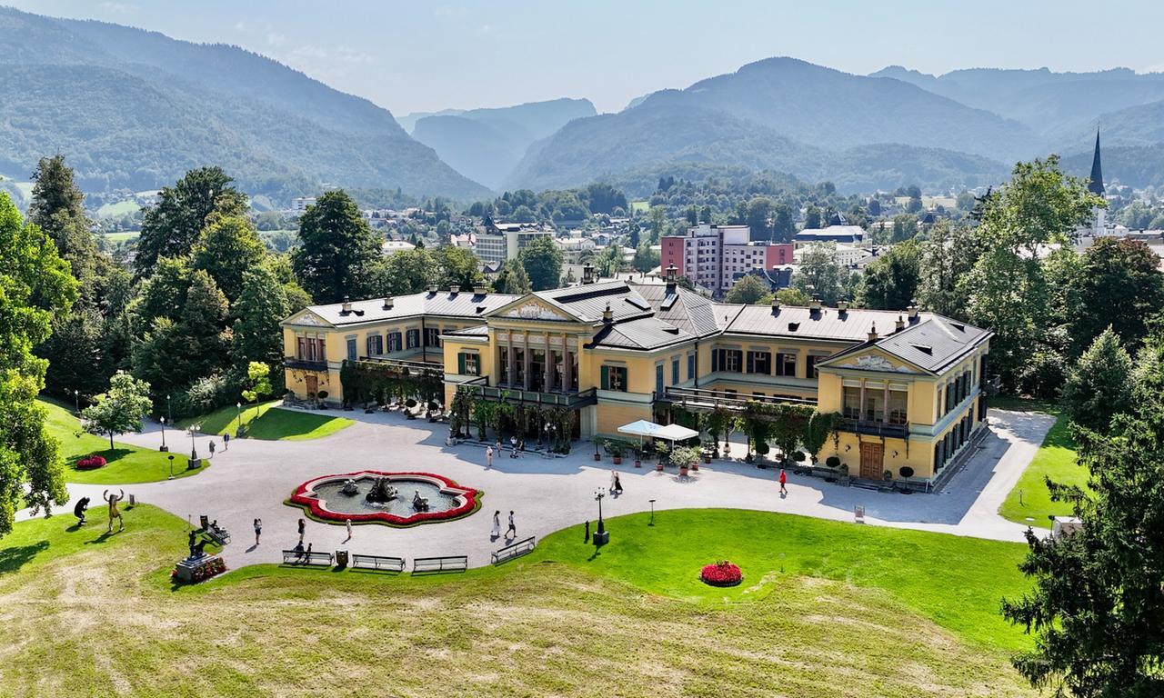 Das Bild zeigt die Kaiservilla in Bad Ischl, Oberösterreich. Die Villa ist ein elegantes, gelb gestrichenes Gebäude mit einem ausgeprägt klassischen Stil und einer breiten, verzierten Fassade. Vor der Villa erstreckt sich ein gepflegter Garten mit kunstvoll angelegten Blumenbeeten, die in Form eines Blütenmotivs angeordnet sind. Im Vordergrund befindet sich ein runder Springbrunnen, umgeben von mehreren Bänken. Im Hintergrund sind sanfte, grüne Hügel und Berge sichtbar, die die Szenerie umrahmen. Es gibt einige Besucher, die durch den Garten spazieren. Das Wetter ist sonnig und klar, was eine freundliche und einladende Atmosphäre schafft. Die Architektur der Umgebung spiegelt den historischen Reichtum und den Glanz der Habsburger-Zeit wider.