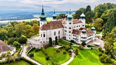 Das Bild zeigt Schloss Artstetten in Niederösterreich. Es handelt sich um ein historisches Bauwerk mit mehreren Türmchen, die in einem charakteristischen, spitzen Design und mit kupferartigen Dächern gestaltet sind. Der Hauptbau des Schlosses ist weiß und hat große Fenster mit grünen Fensterrahmen. Der Schlossbereich ist von gepflegten Gärten und Grünanlagen umgeben, in denen Sträucher und kleine Bäume zu sehen sind. Mehrere Wege schlängeln sich durch die Gartenlandschaft. Im Vordergrund des Bildes sind einige Menschen, die sich in der Gartenanlage aufhalten. Im Hintergrund sind sanfte Hügel und Bäume sichtbar, die die landschaftliche Umgebung des Schlosses prägen. Der Himmel ist bewölkt, was dem Bild eine ruhige und friedliche Atmosphäre verleiht.