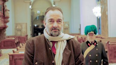 Ein Mann mit einem Bart und einem beigen Schal steht in einer beleuchteten Kirche, hinter ihm ist ein Soldat in historischer Uniform zu sehen.