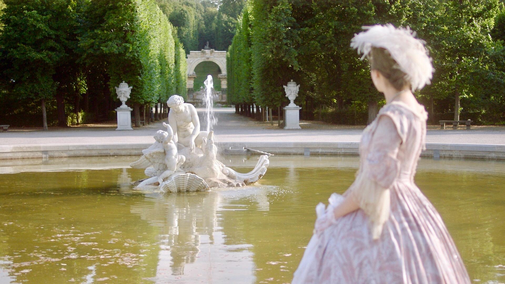 Frau in historischem Kleid blickt auf eine Brunnenfigur in den Gartenanlagen von Schloss Schönbrunn, im Hintergrund ein von Bäumen gesäumter Weg.