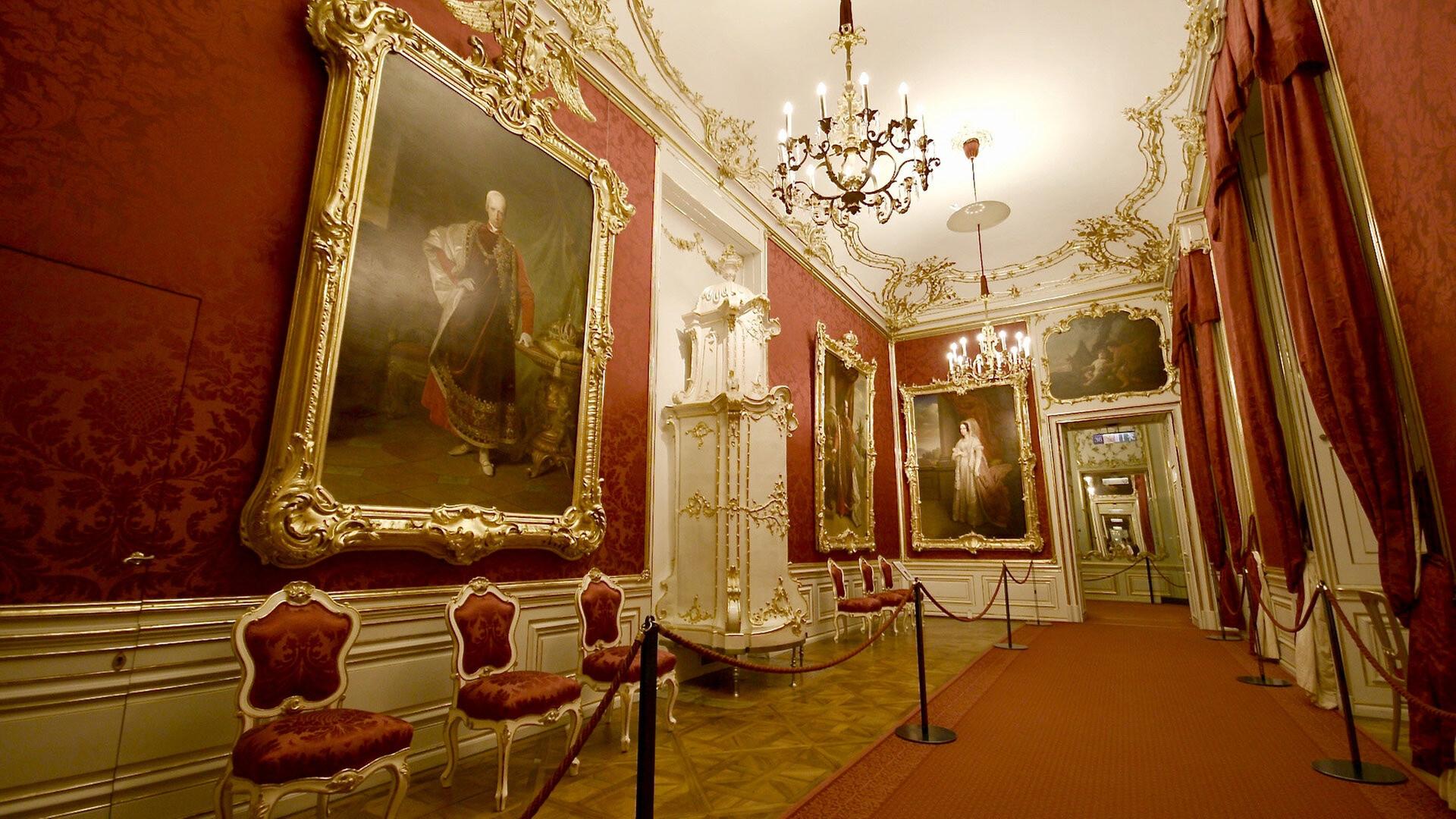 Prunkvoll dekorierter Rokokosaal mit roten Wänden, vergoldeten Gemälden, Stuckverzierungen, Kristallleuchtern und antiken Stühlen, abgesperrt mit roten Samtseilen.