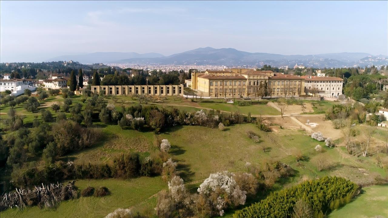 Villa Medici Imperiale in Florenz, umgeben von weitläufigen Grünflächen und Bäumen, im Hintergrund die Stadt Florenz und hügelige Landschaft.